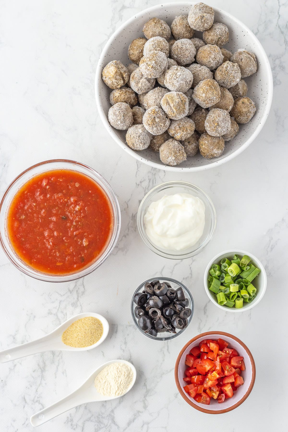 Bowls of frozen meatballs, enchilada sauce, sour cream, sliced black olives, diced tomatoes, chopped green onions, and two spoons with powdered seasonings on a white marble surface.