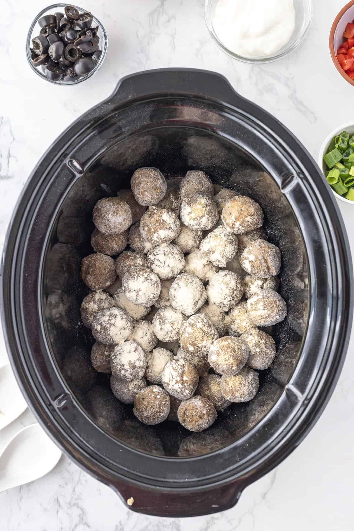 A slow cooker filled with frozen meatballs coated in white seasoning, surrounded by small bowls of sliced olives, sour cream, green onions, and diced tomatoes.