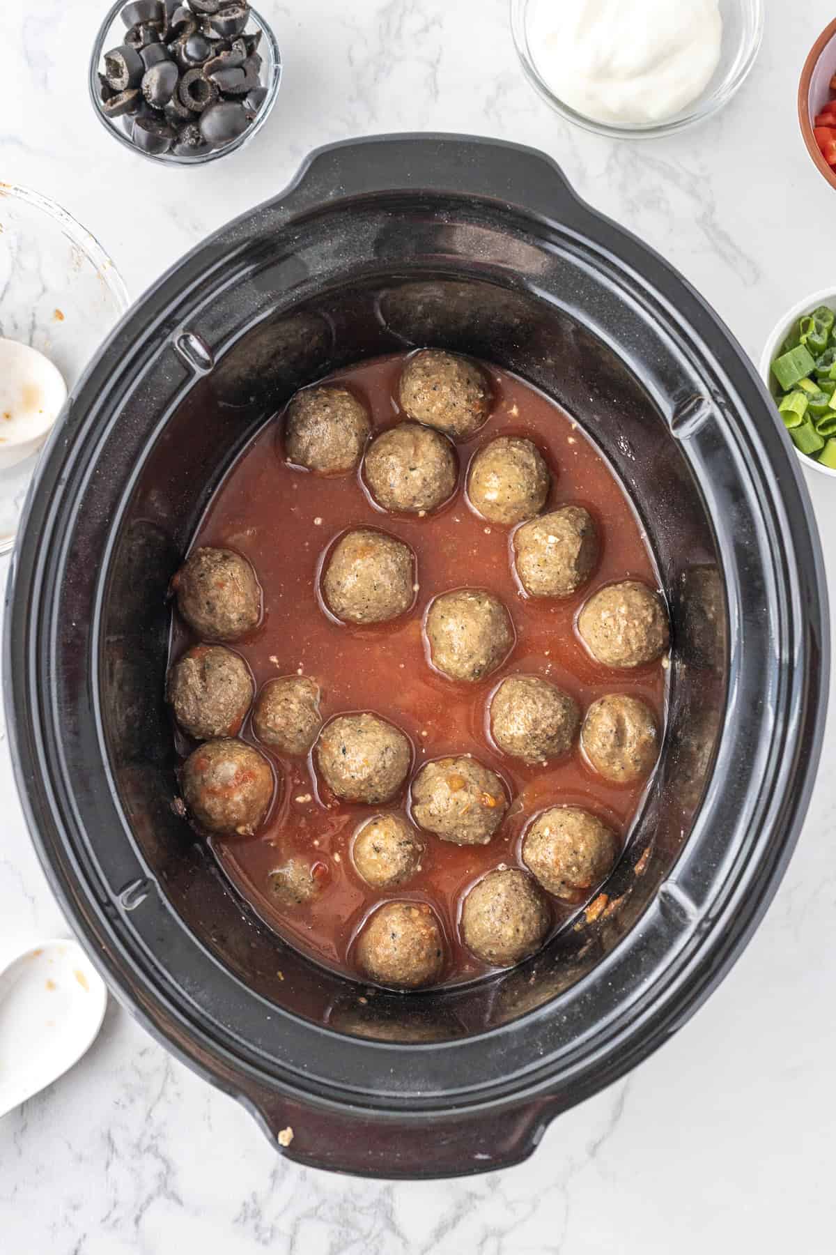 A slow cooker filled with meatballs in enchilada sauce, surrounded by small bowls of sliced olives, green onions, sour cream, and diced tomatoes.