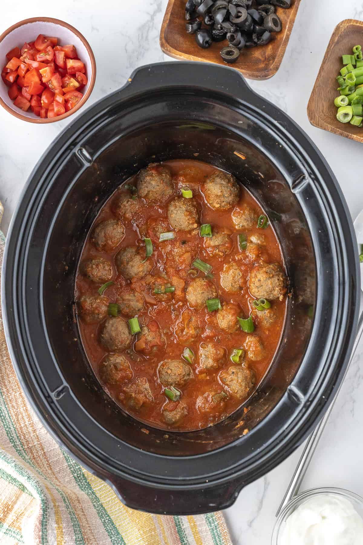 Meatballs in enchilada sauce garnished with chopped green onion in a slow cooker, surrounded by bowls of diced tomatoes, sliced black olives, and green onions.