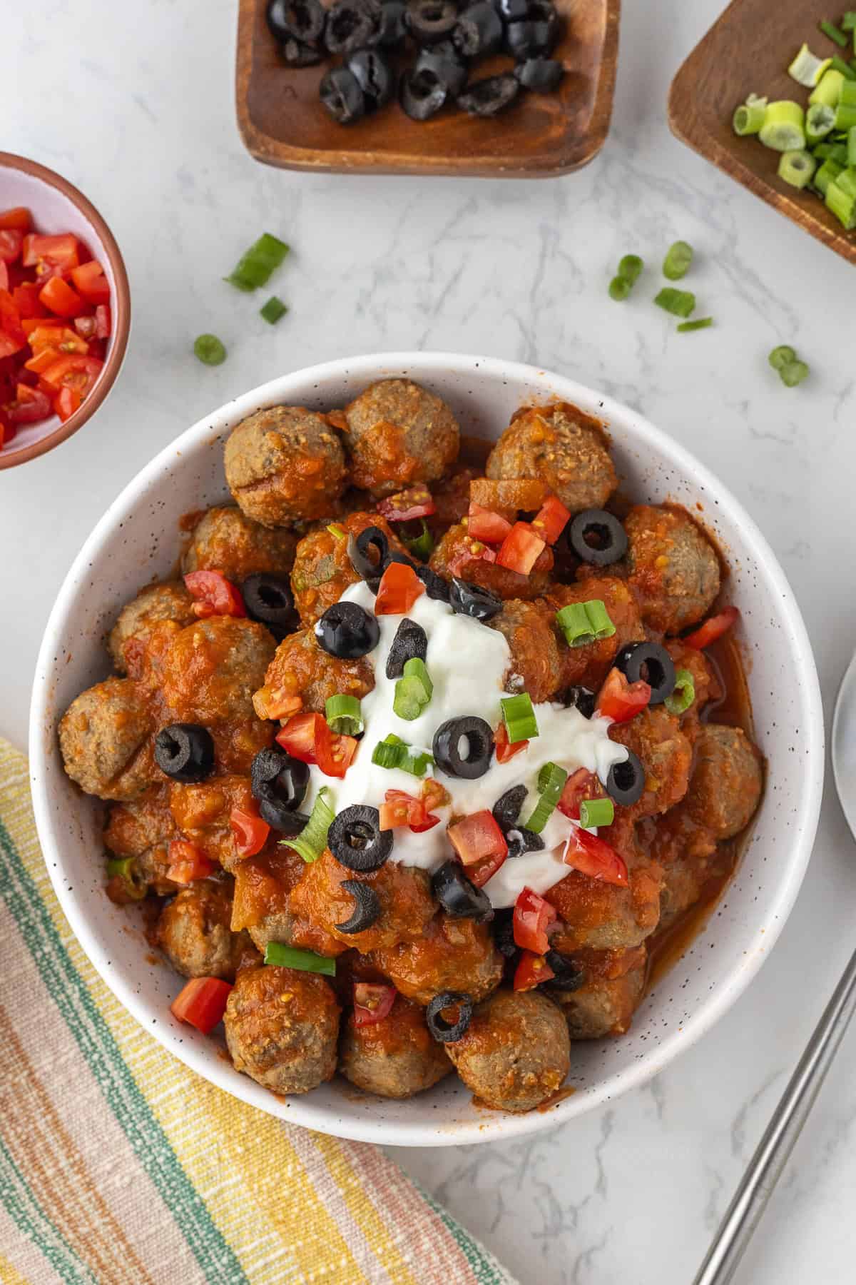 A bowl of meatballs topped with enchilada sauce, sour cream, chopped red bell peppers, sliced black olives, and green onions on a white surface.