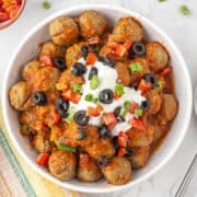 A bowl of meatballs topped with enchilada sauce, sour cream, sliced black olives, chopped tomatoes, and green onions on a marble surface.