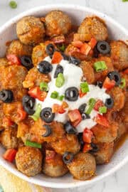 A bowl of meatballs topped with enchilada sauce, sour cream, sliced black olives, chopped tomatoes, and green onions on a marble surface.