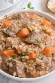 A bowl of beef and barley stew with chunks of beef, carrots, and barley in a thick broth, garnished with chopped parsley. Slices of bread are visible in the background.