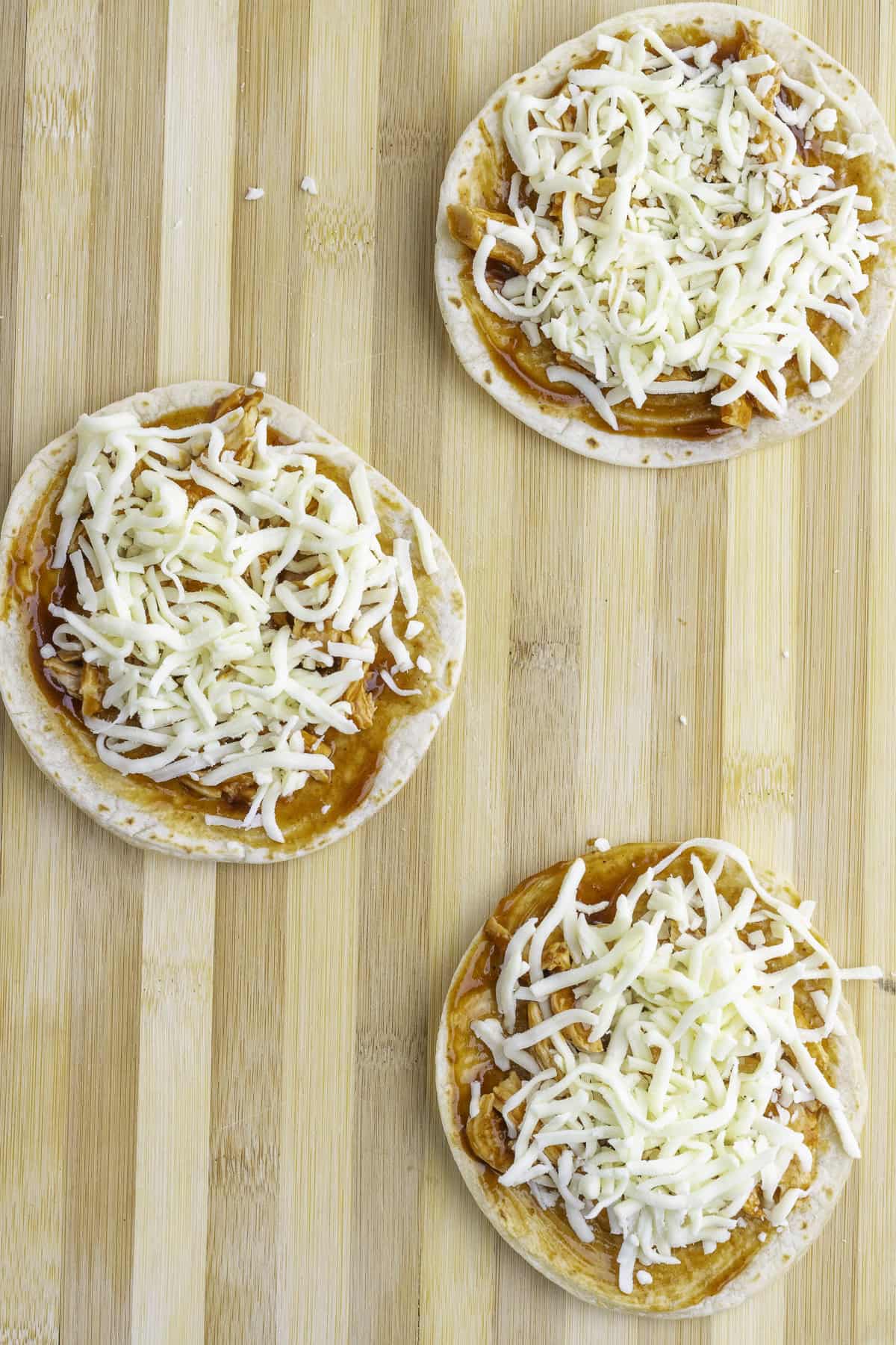 Three small round pizza crusts topped with tomato sauce and shredded mozzarella cheese are arranged on a wooden surface.