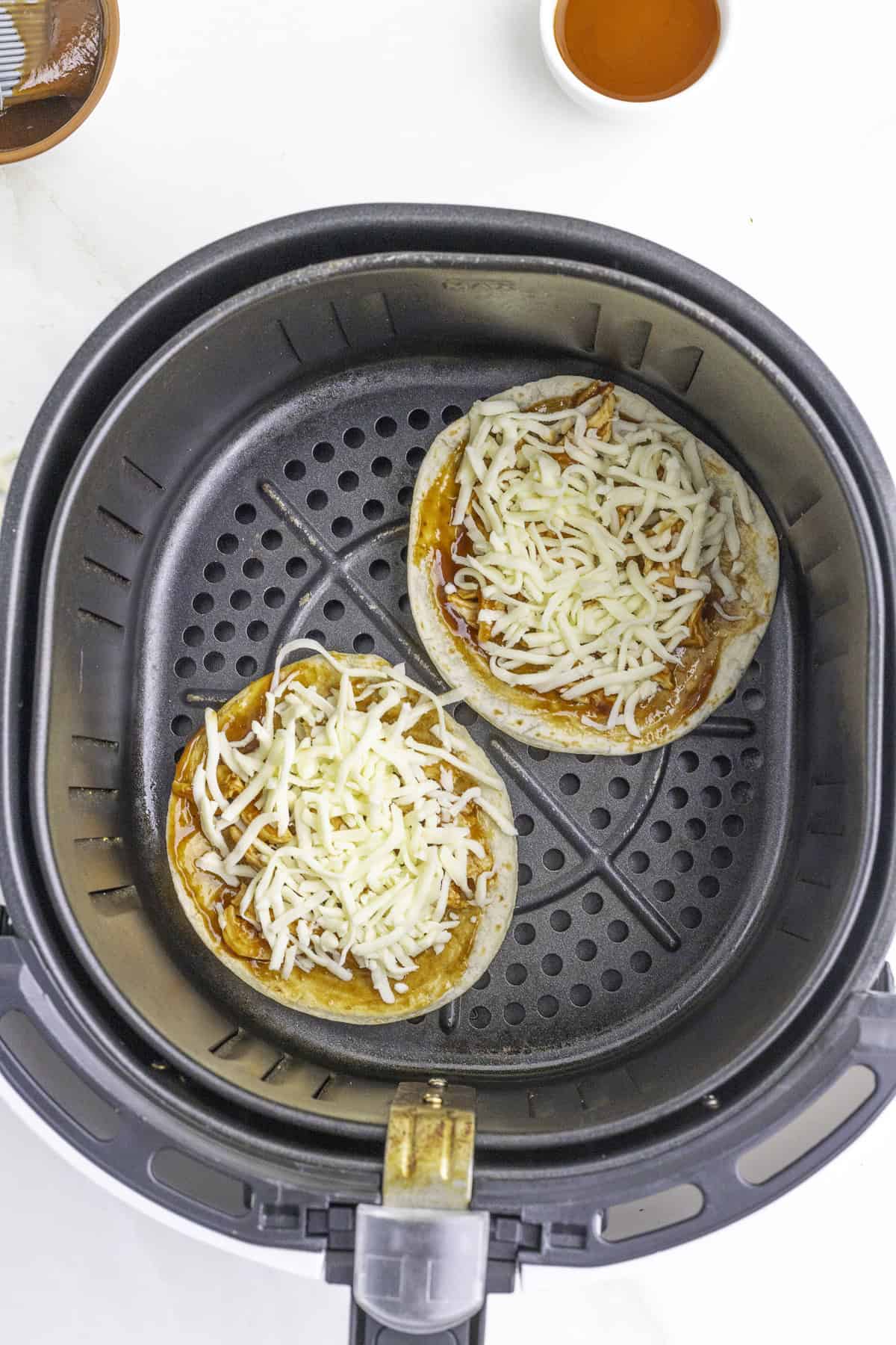 Two English muffin halves topped with sauce and shredded cheese are placed in an air fryer basket, ready to be cooked.