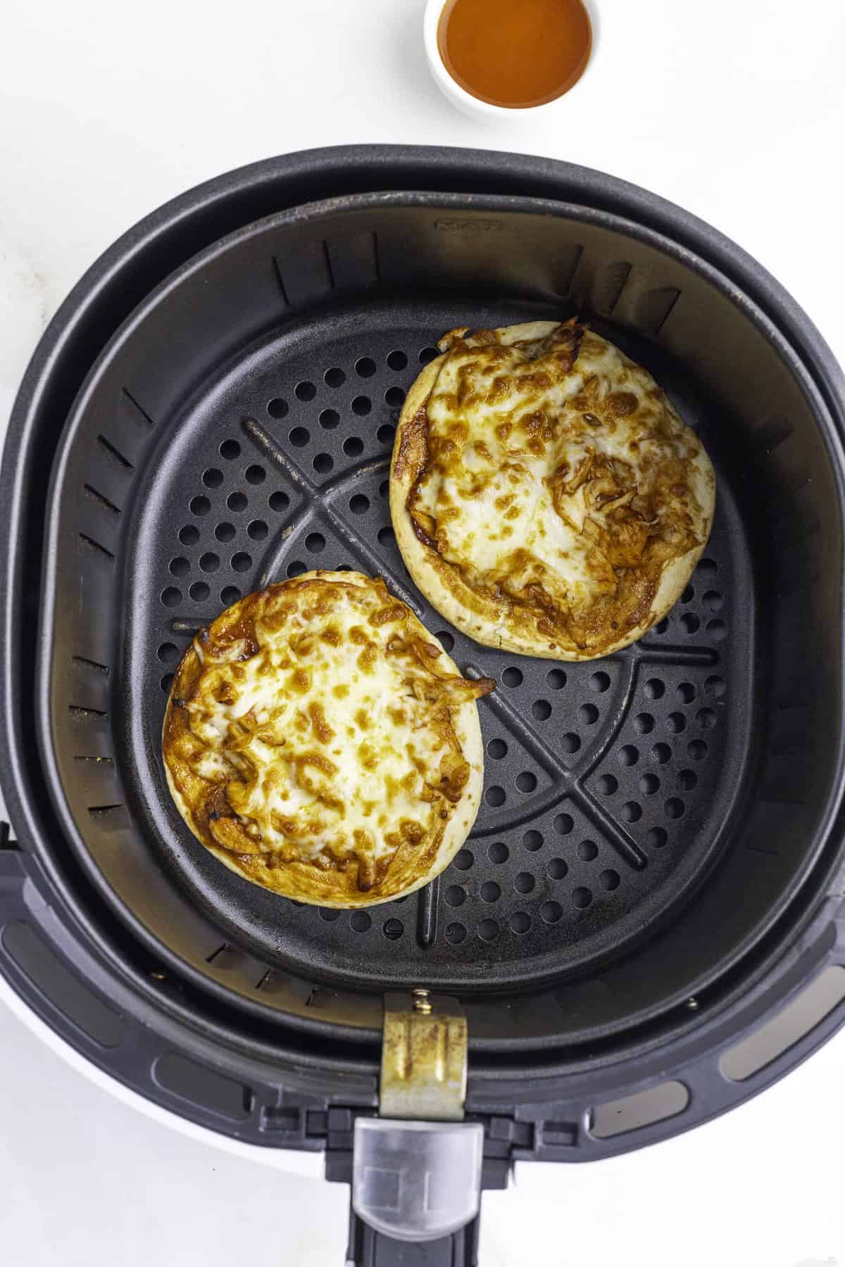 Two mini pizzas with melted cheese and browned edges inside an air fryer basket, with a small bowl of orange sauce in the background.