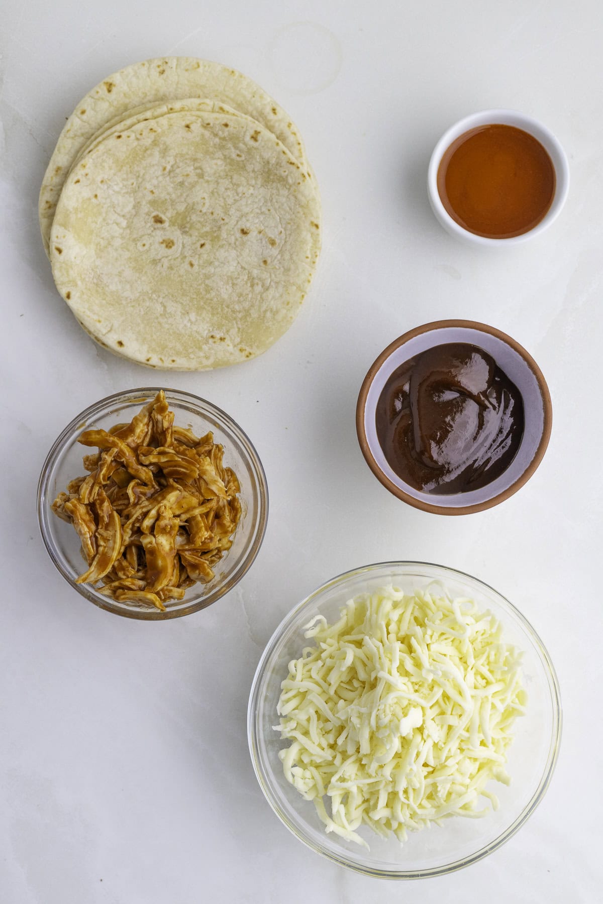 Tortillas, shredded chicken, barbecue sauce, shredded cheese, and hot sauce are arranged separately on a white surface.