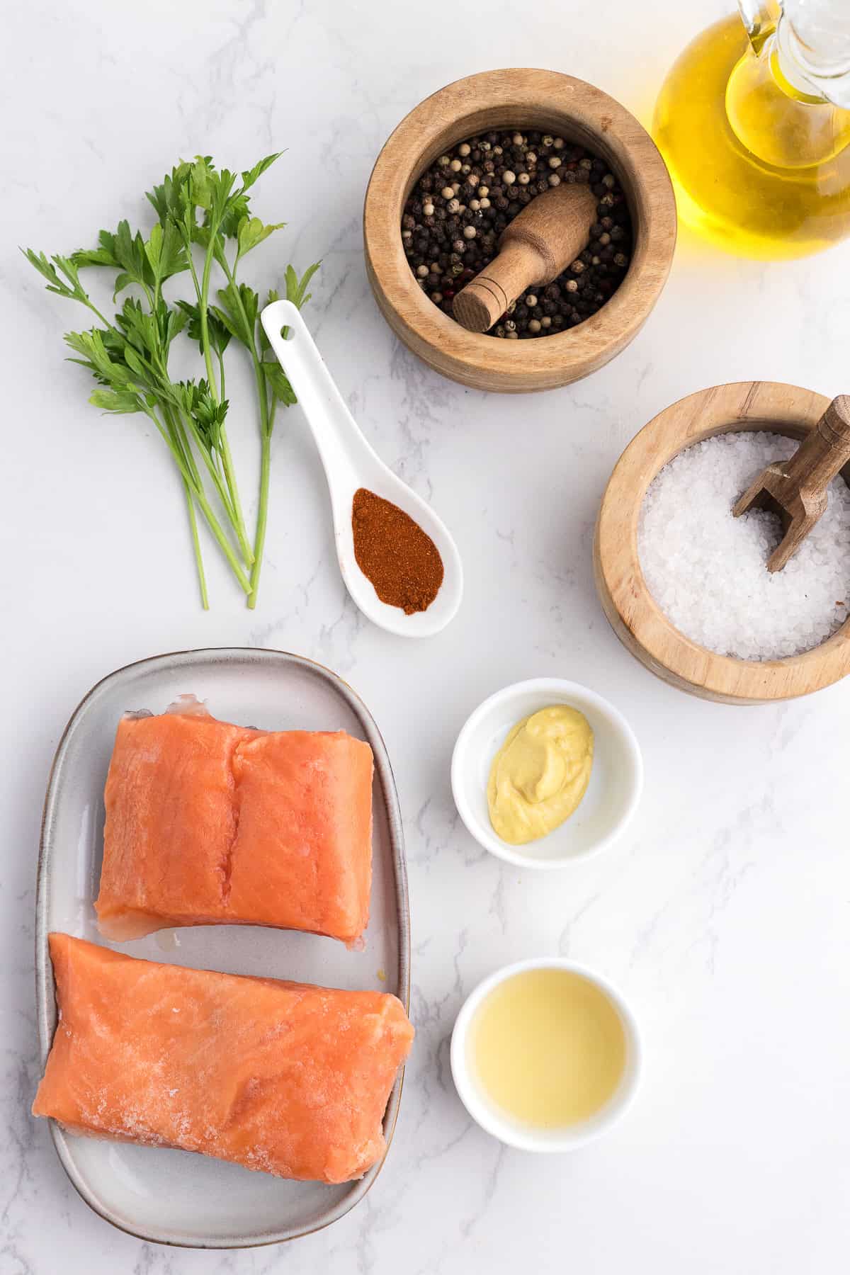 Deux filets de saumon crus sur une assiette avec du persil, du poivre noir, de l'huile d'olive, du sel, de la moutarde, du vinaigre et du paprika disposés sur une surface en marbre blanc.