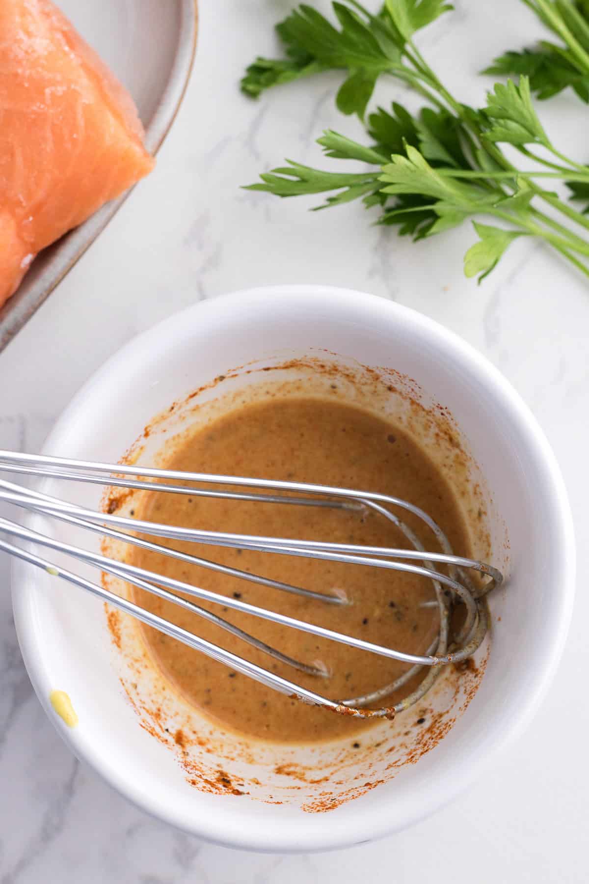 Un fouet en métal dans un bol blanc contenant une marinade brune et assaisonnée. Du persil frais et une assiette contenant du saumon se trouvent à proximité, sur une surface claire.
