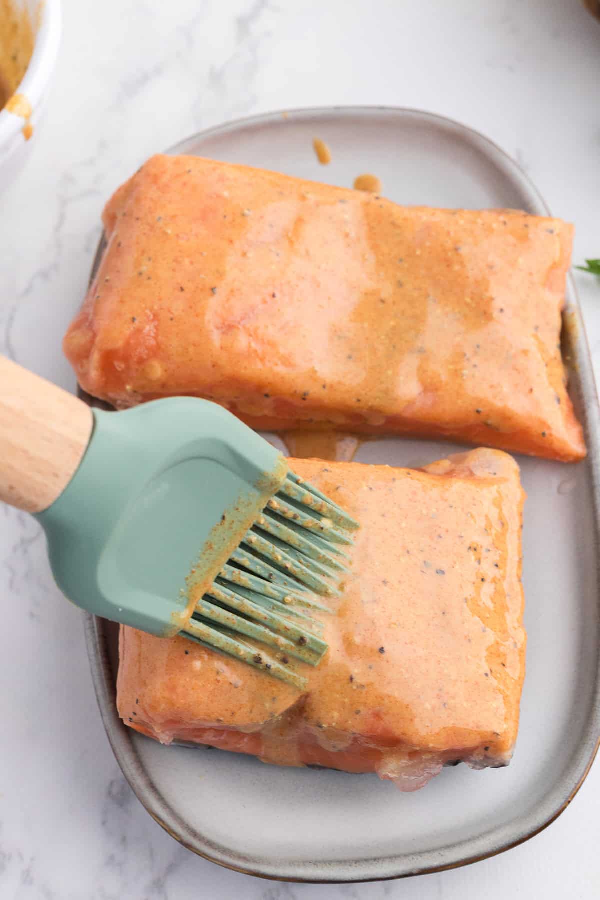 Deux filets de saumon sur une assiette, badigeonnés d'une marinade orange à l'aide d'un pinceau en silicone vert.