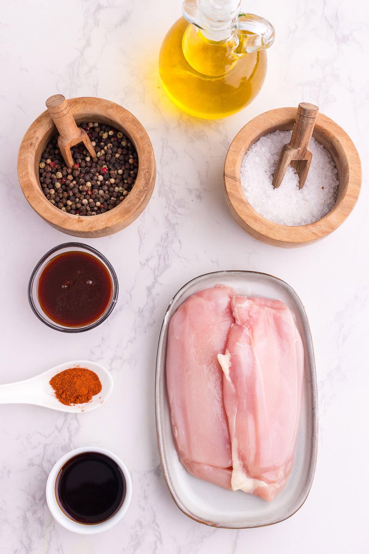 Raw chicken breasts on a plate, surrounded by bowls of peppercorns, salt, olive oil, BBQ Sauce, soy sauce and a spoon of paprika, all arranged on a white marble surface.