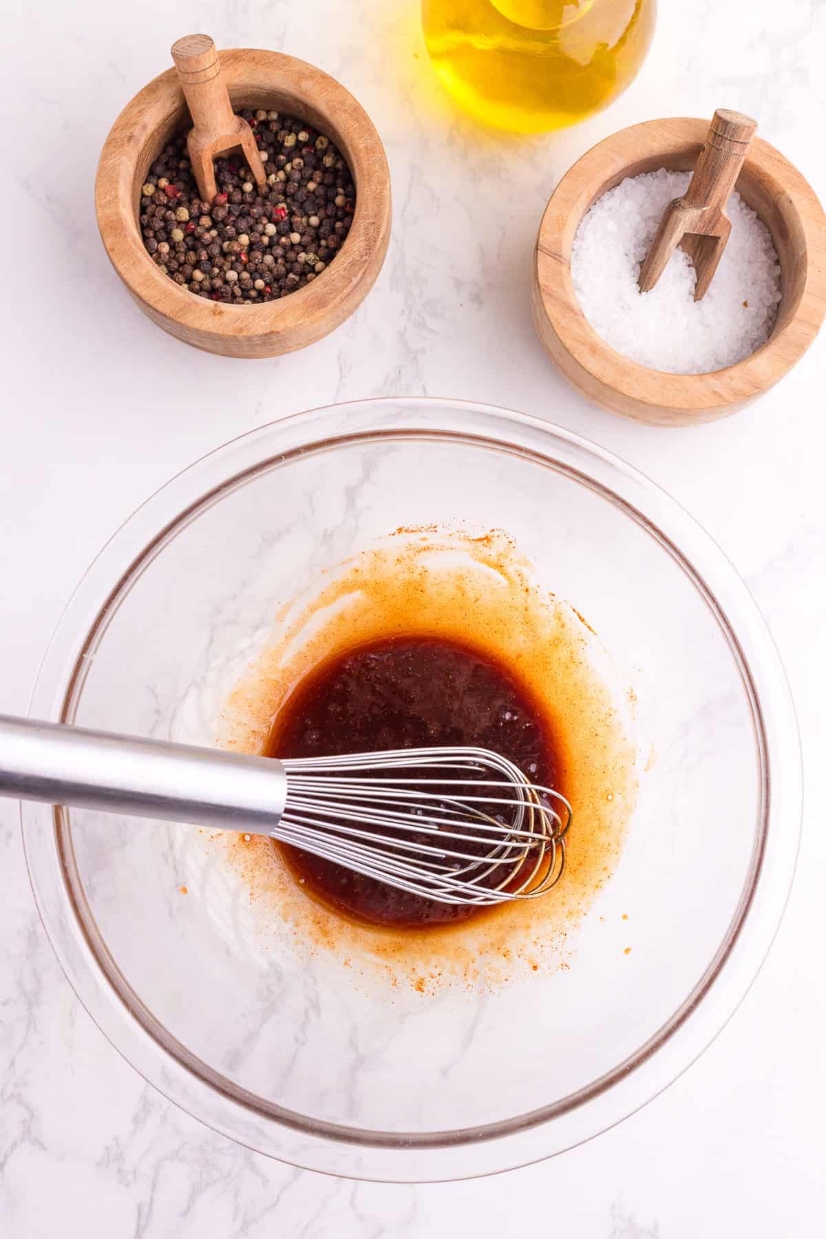 A whisk in a glass bowl mixing a brown liquid, with containers of black pepper, salt, and a bottle of oil on a white surface.