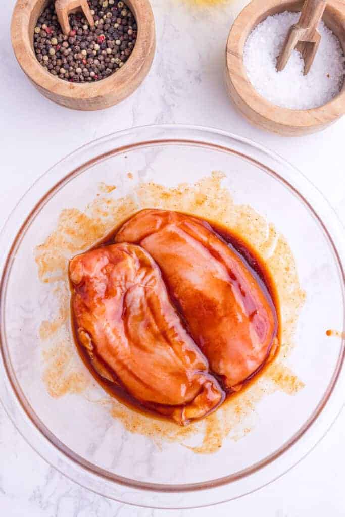 Two raw chicken breasts coated in marinade sit in a glass bowl on a white surface, with bowls of black pepper and salt nearby.