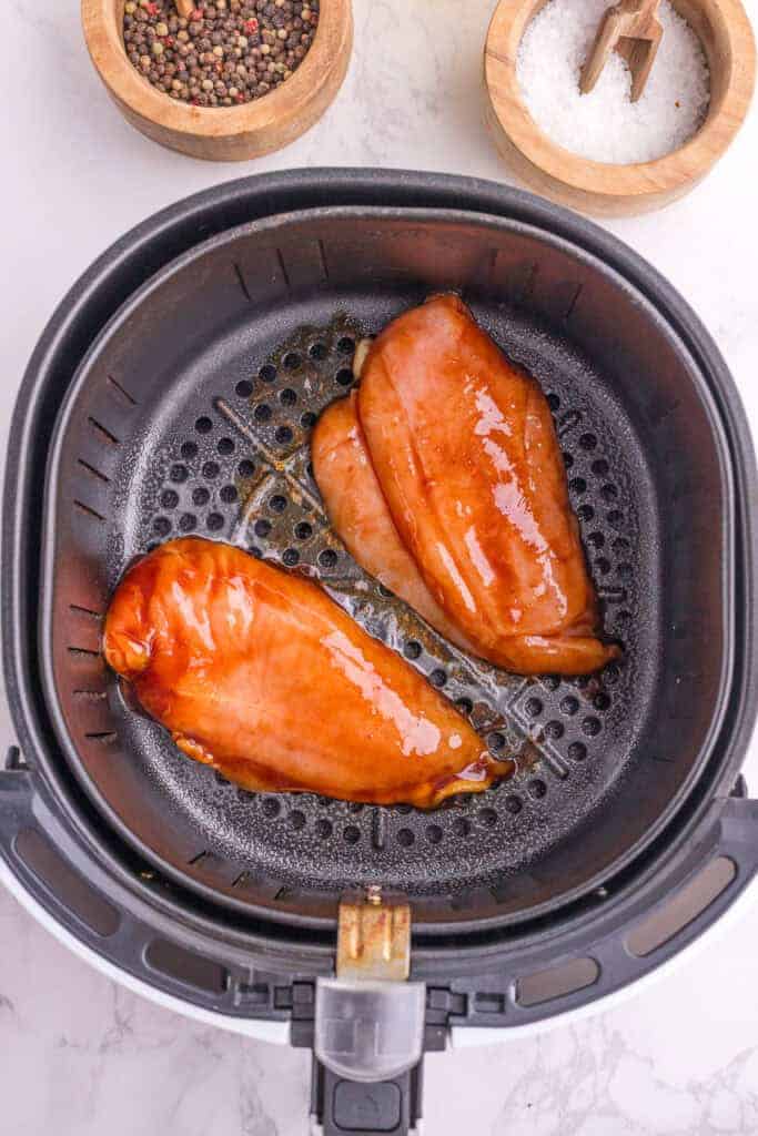 Two raw, glazed chicken breasts placed inside an air fryer basket, with bowls of peppercorns and salt in the background.