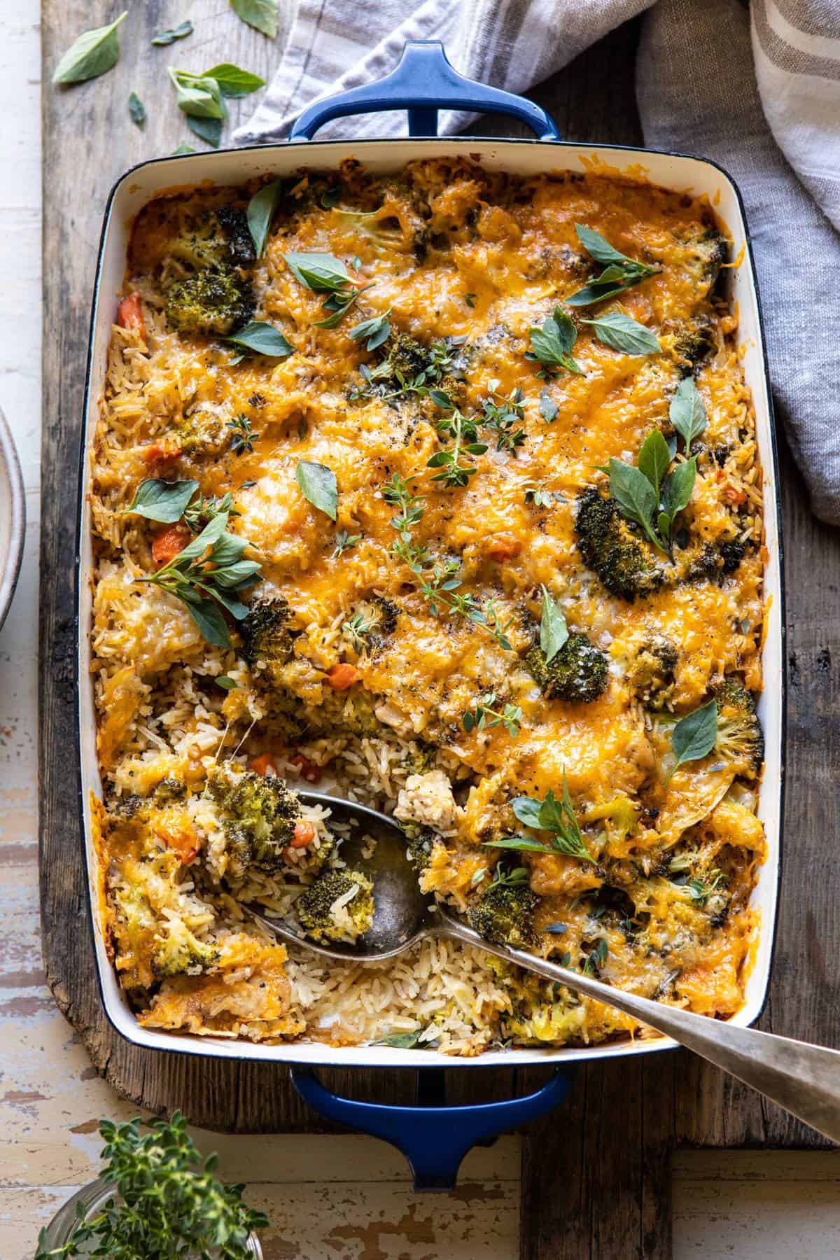 A baked casserole dish filled with rice, broccoli, vegetables, and melted cheese, garnished with fresh herbs, with a serving spoon placed inside.