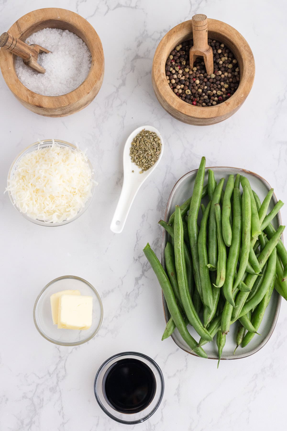 Une assiette de haricots verts frais, de fromage râpé, de beurre, de vinaigre balsamique, de sel, de poivre et d'herbes séchées disposés sur une surface de marbre blanc.