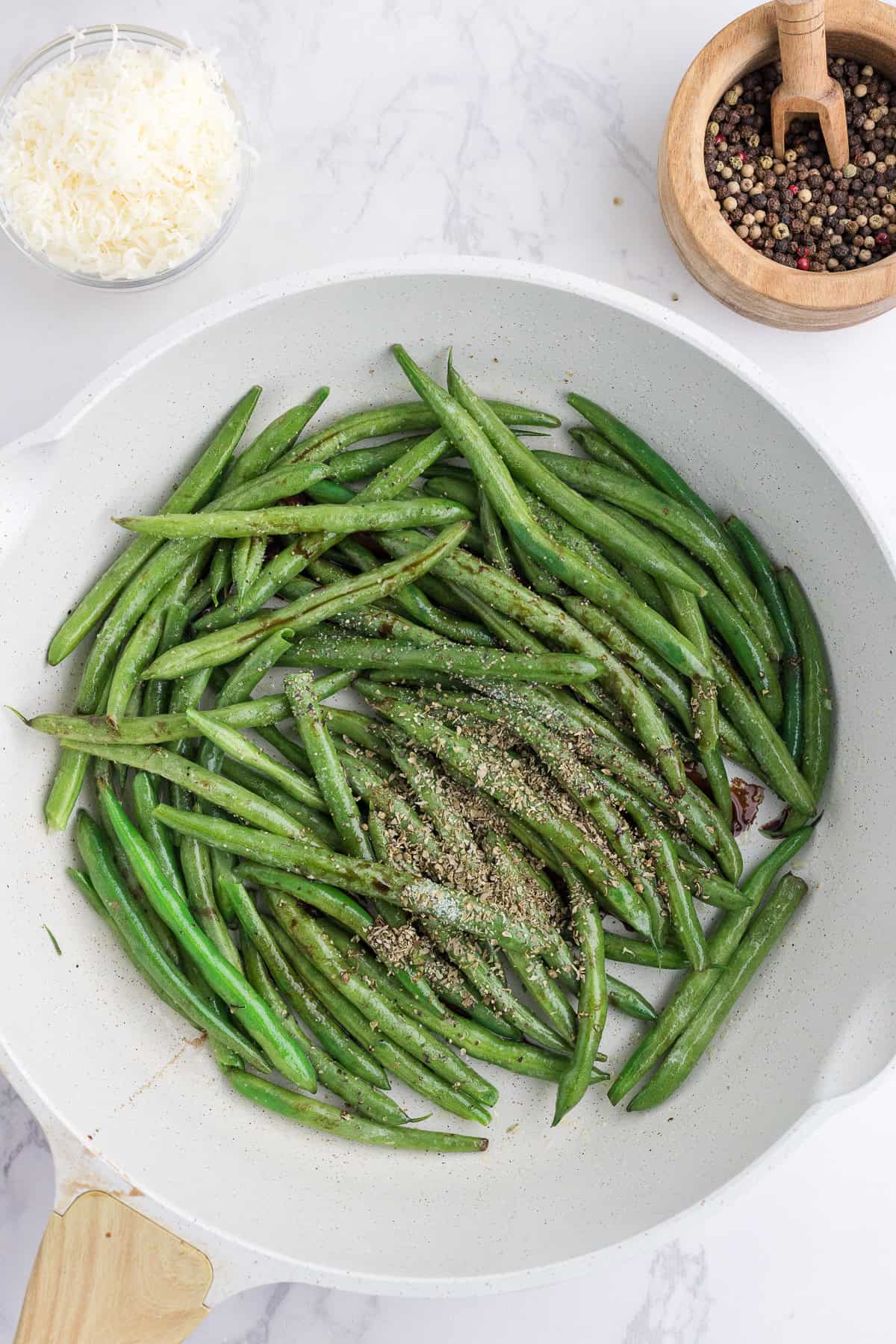 Haricots verts cuits dans une poêle blanche, assaisonnés de poivre, avec un bol de fromage râpé et un moulin à poivre en bois à proximité sur une surface en marbre.