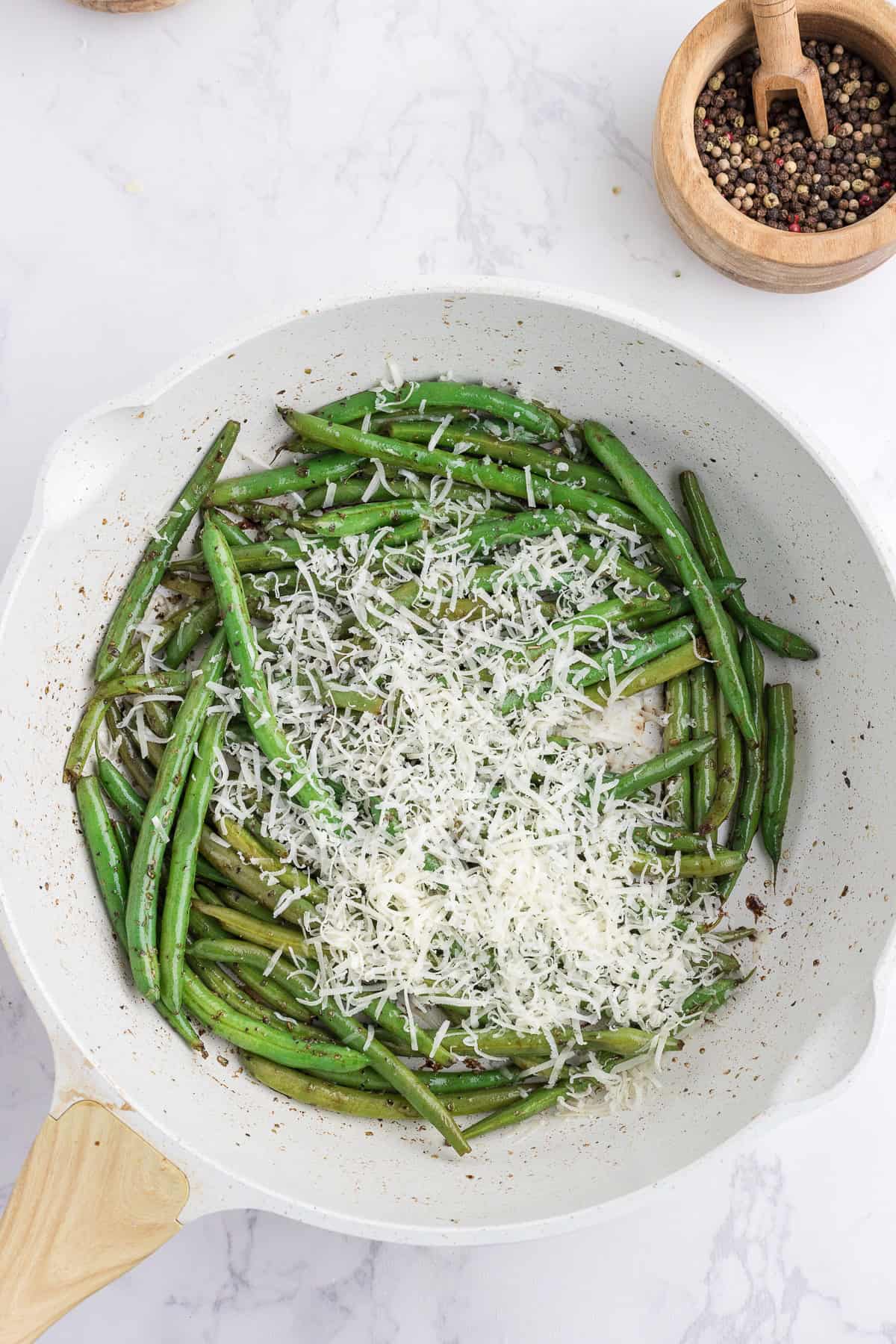 Haricots verts sautés dans une poêle blanche, garnis de fromage râpé. Un bol de grains de poivre mélangés est visible à l'arrière-plan sur une surface en marbre.