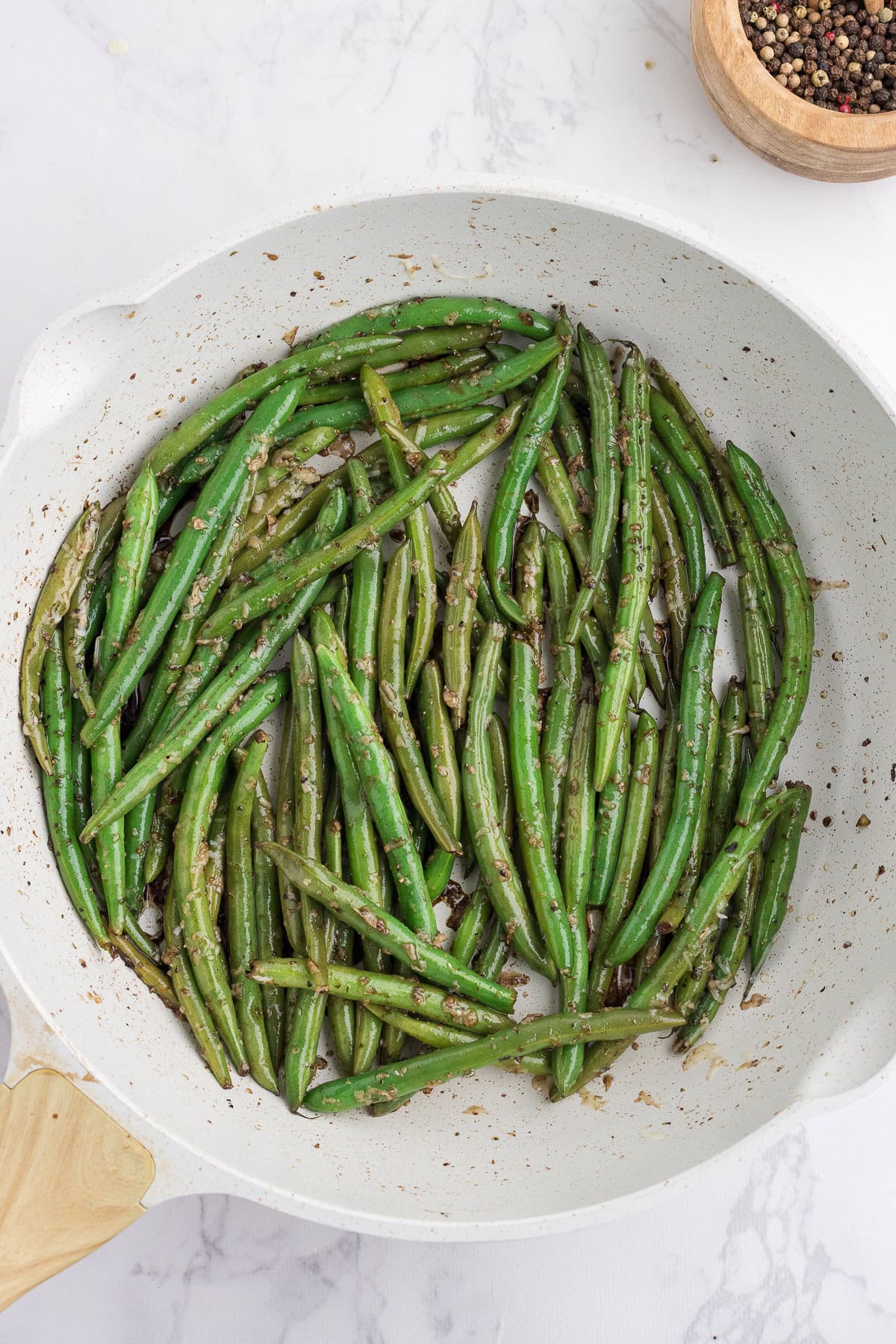 Une poêle blanche contenant des haricots verts cuits et assaisonnés d'herbes et d'épices, posée sur une surface en marbre.