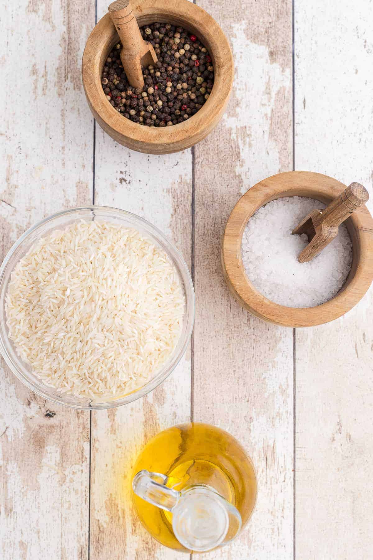 A bowl of dry basmati rice, a bowl of salt, a bowl of peppercorns, and a glass container of oil arranged on a white wooden surface.