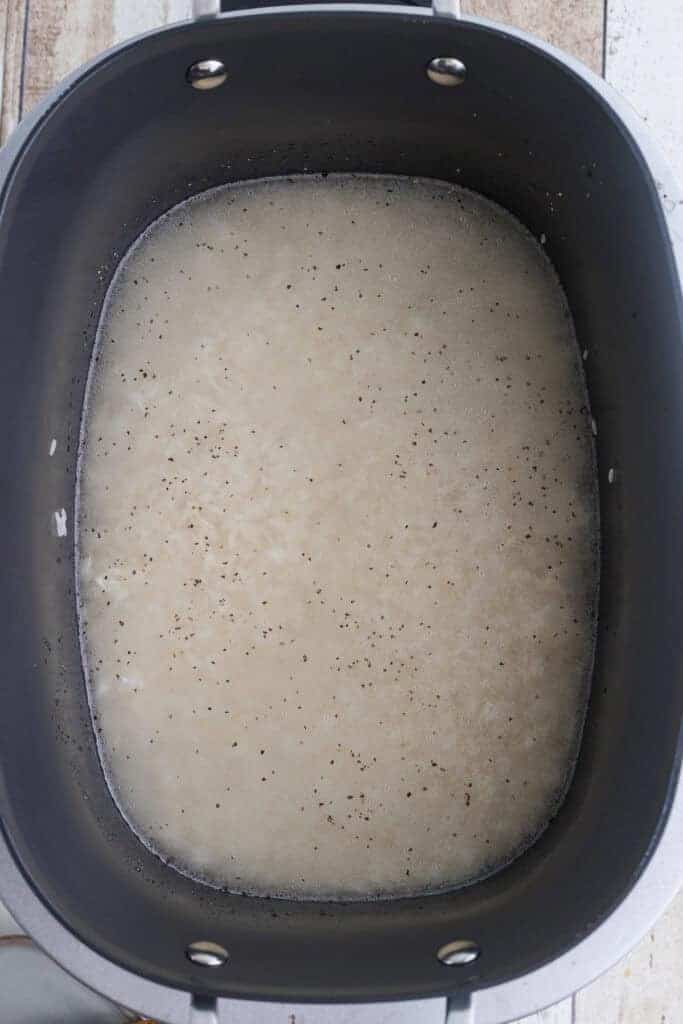 A black pot filled with uncooked rice, water, and visible black pepper, sitting on a light-colored surface.
