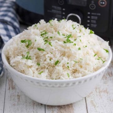 A white bowl filled with cooked white rice, garnished with chopped parsley, sits on a light wooden surface near a blue-striped cloth.