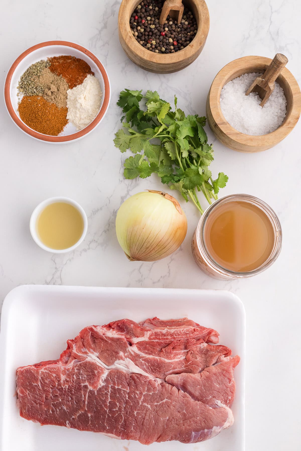 Raw beef on a tray, whole onion, fresh cilantro, spice mix, salt, peppercorns, lime juice, and broth arranged on a white countertop.