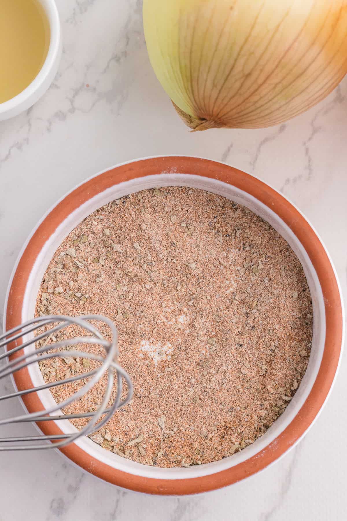 A bowl of dry seasoning mix with a metal whisk beside it, an onion, and a small bowl of liquid on a marble countertop.