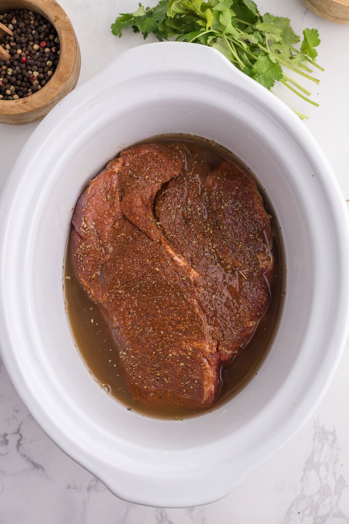 Raw, seasoned beef inside a white slow cooker, surrounded by a bowl of peppercorns and a bunch of fresh cilantro on a marble countertop.