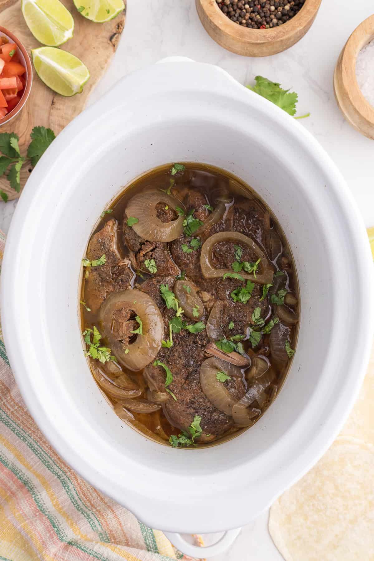 A slow cooker filled with cooked beef, sliced onions, and chopped cilantro, surrounded by lime wedges, peppercorns, and fresh cilantro on a countertop.