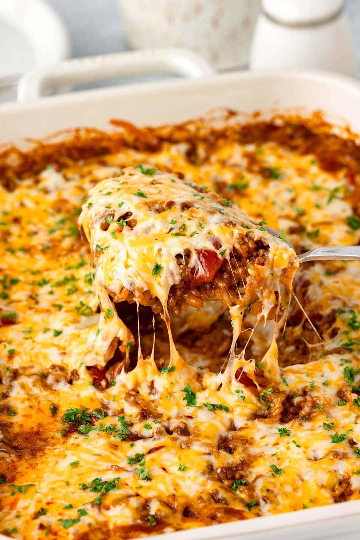 A serving spoon lifts a portion of cheesy casserole topped with melted cheese and herbs from a baking dish.