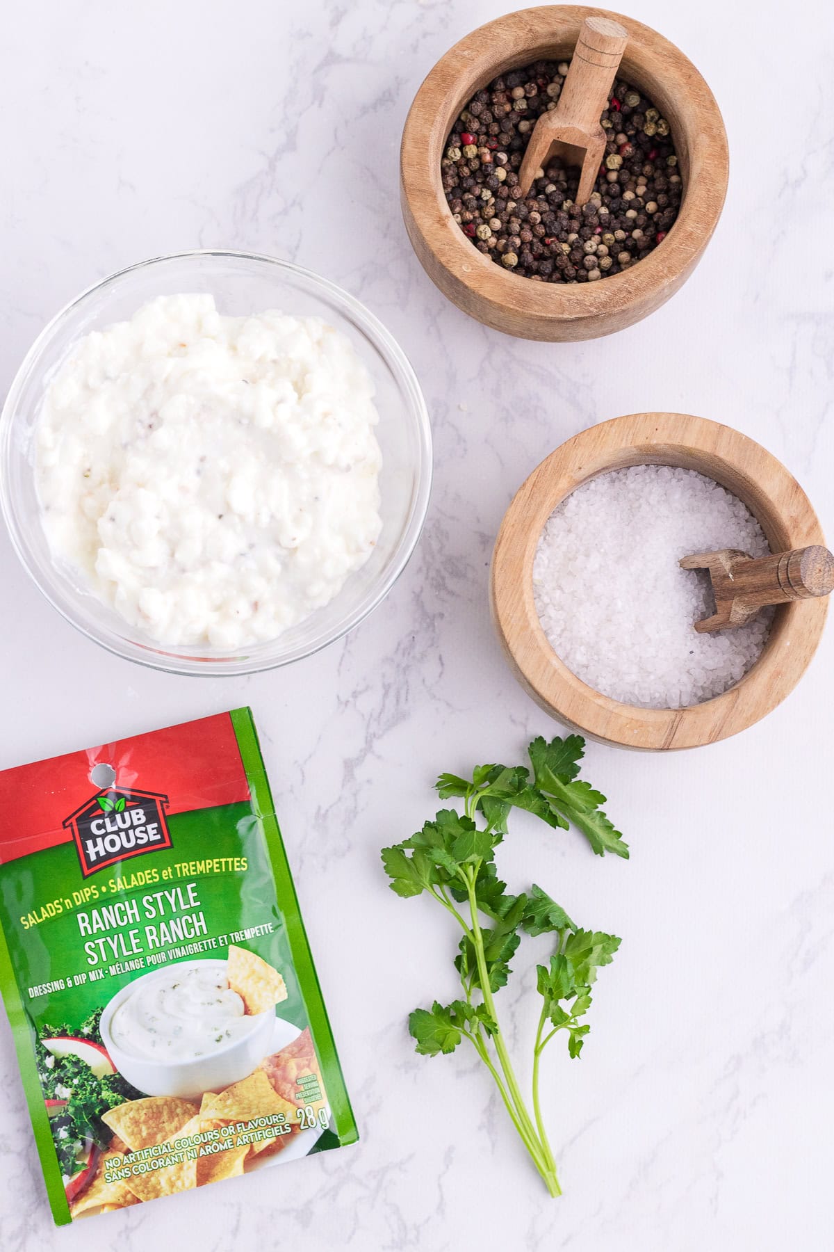 A bowl of cottage cheese, a bowl of mixed peppercorns, a bowl of salt, a Ranch seasoning packet, and a sprig of parsley on a marble surface.