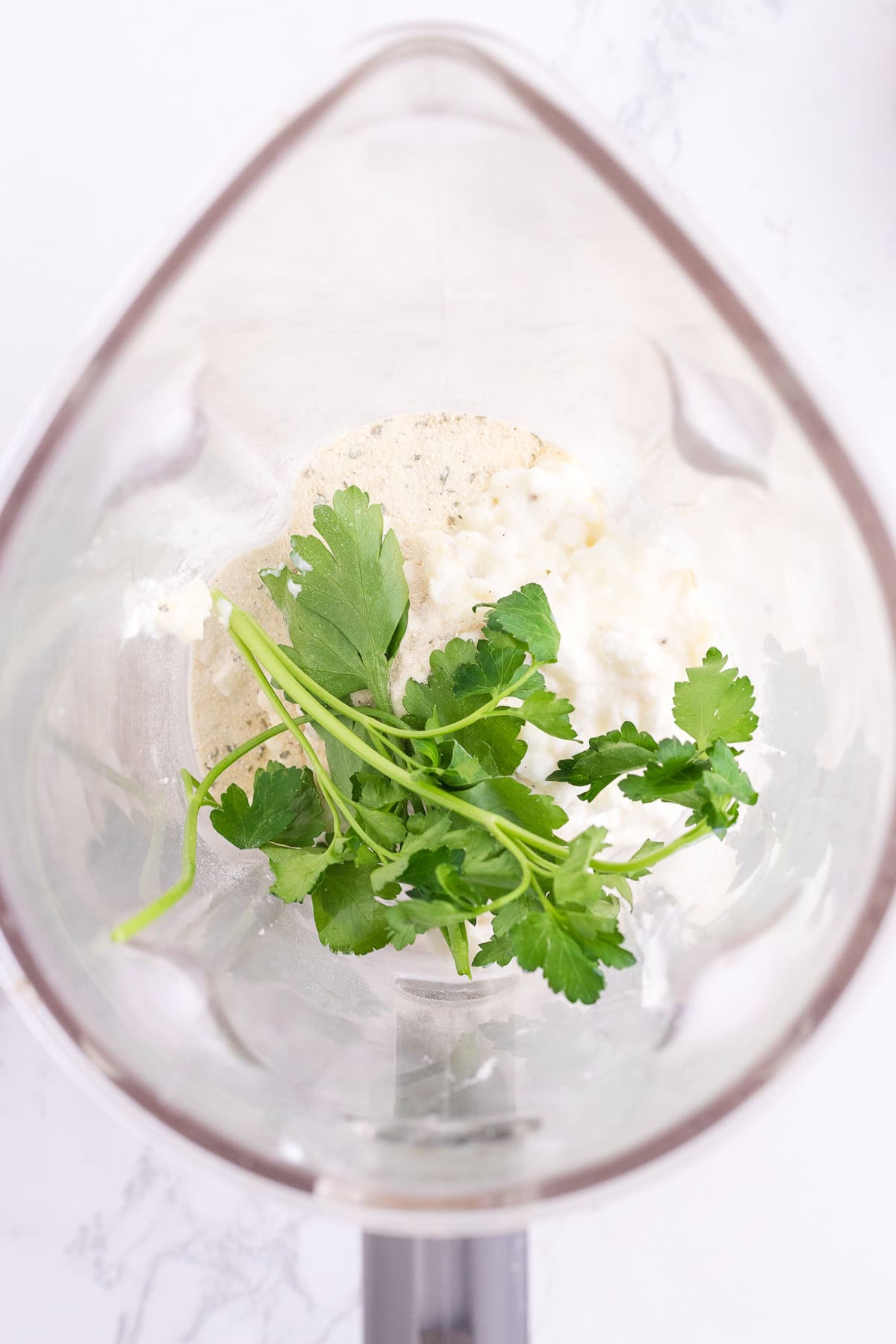 Overhead view of a blender containing fresh parsley, cottage cheese, and ranch seasonning on a white surface.