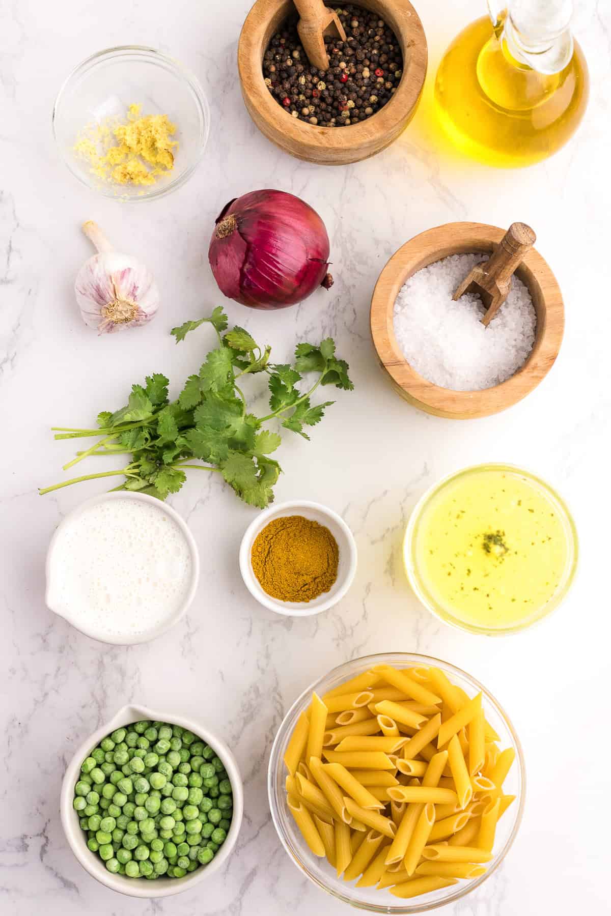 Ingredients on a white surface: penne pasta, green peas, coconut milk, turmeric, cilantro, garlic, red onion, black pepper, sea salt, olive oil, lemon zest, and yellow sauce.