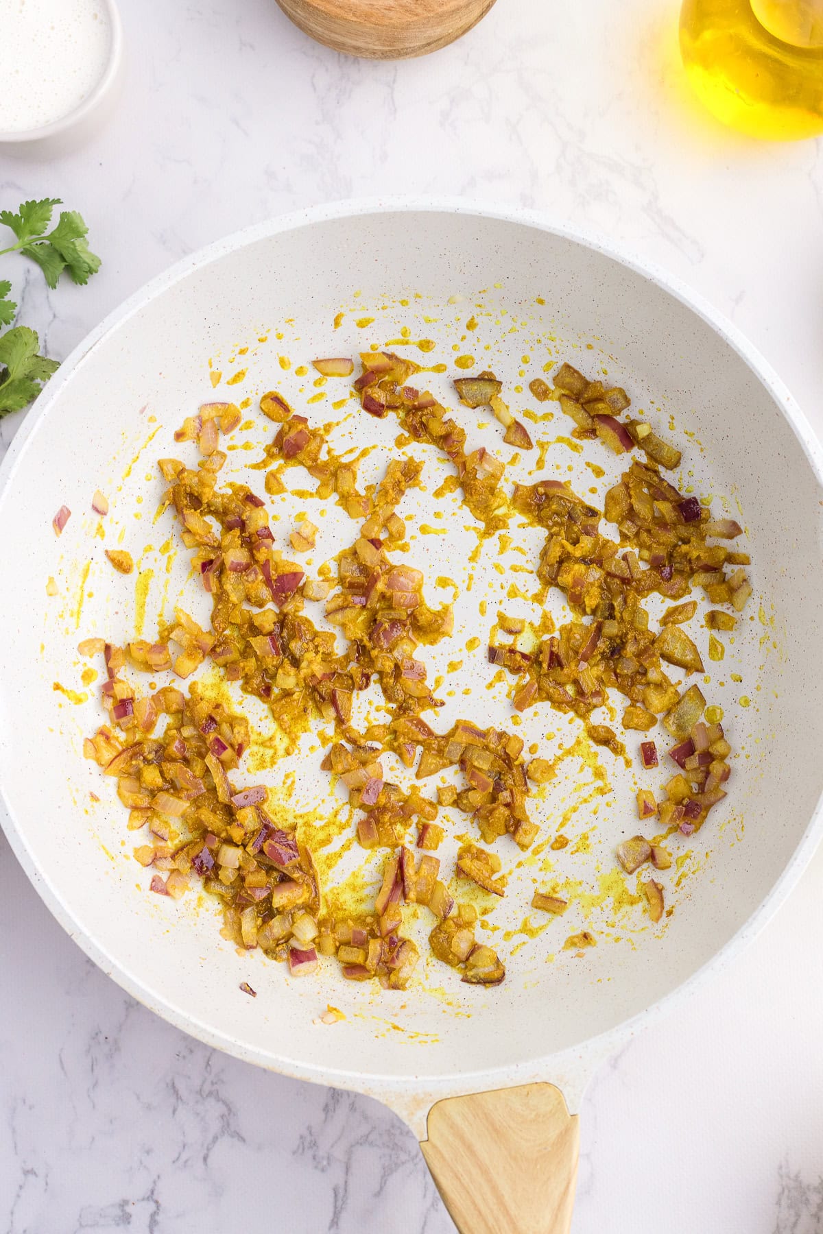 Chopped onions sautรฉing with yellow spices in a white frying pan on a marble countertop.