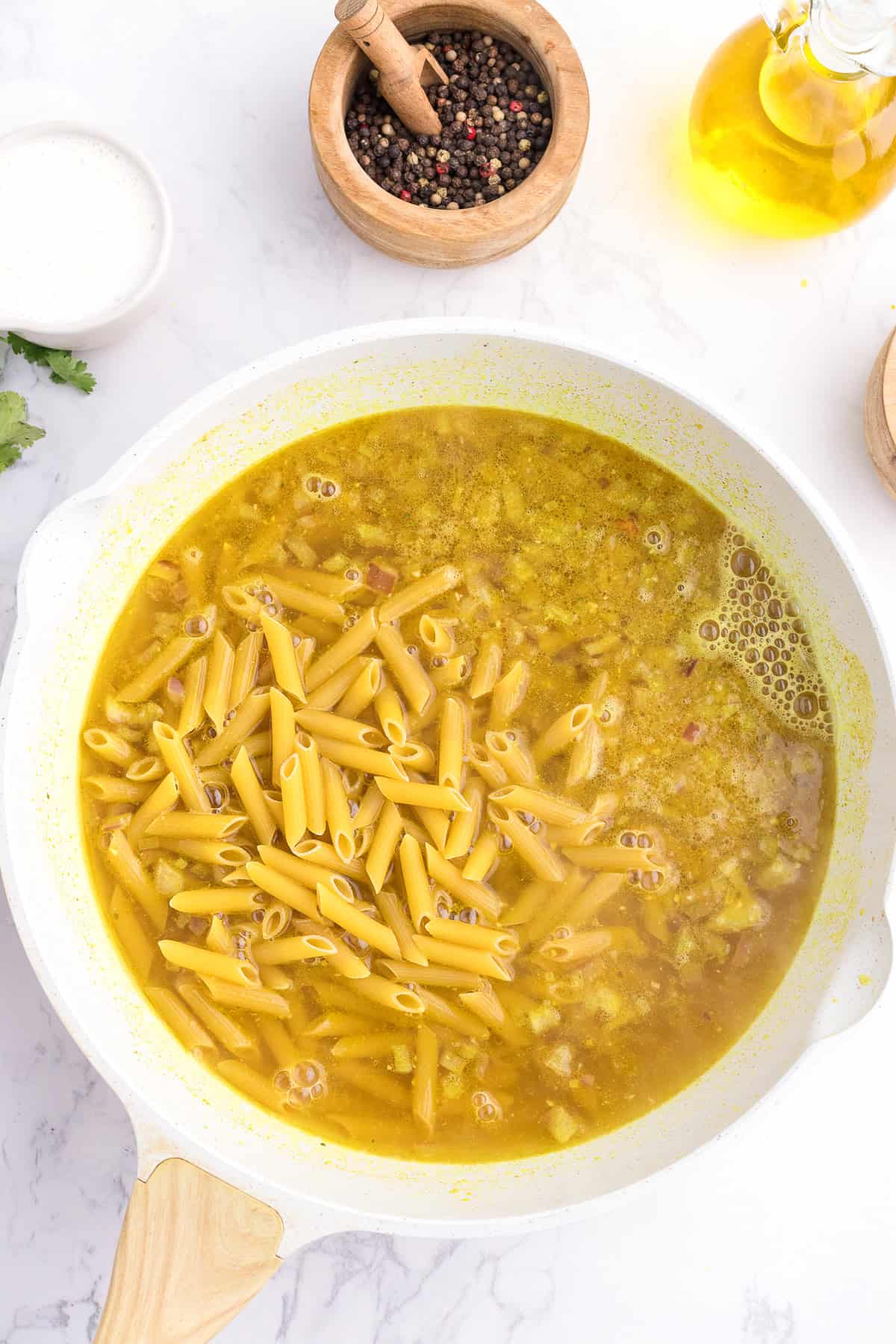 Uncooked penne pasta in a yellow broth inside a white skillet, surrounded by a bowl of peppercorns, olive oil, and herbs on a white countertop.