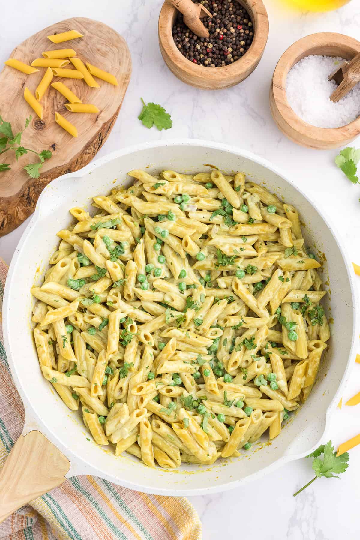 A skillet filled with creamy penne pasta and green peas, surrounded by bowls of peppercorns, salt, uncooked pasta, and fresh herbs on a marble surface.