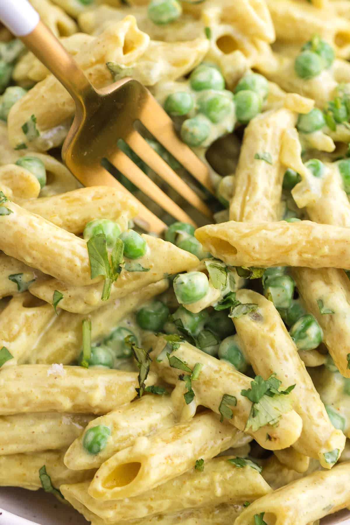 Close-up of penne pasta mixed with green peas and creamy sauce, garnished with chopped herbs, with a fork partially visible.