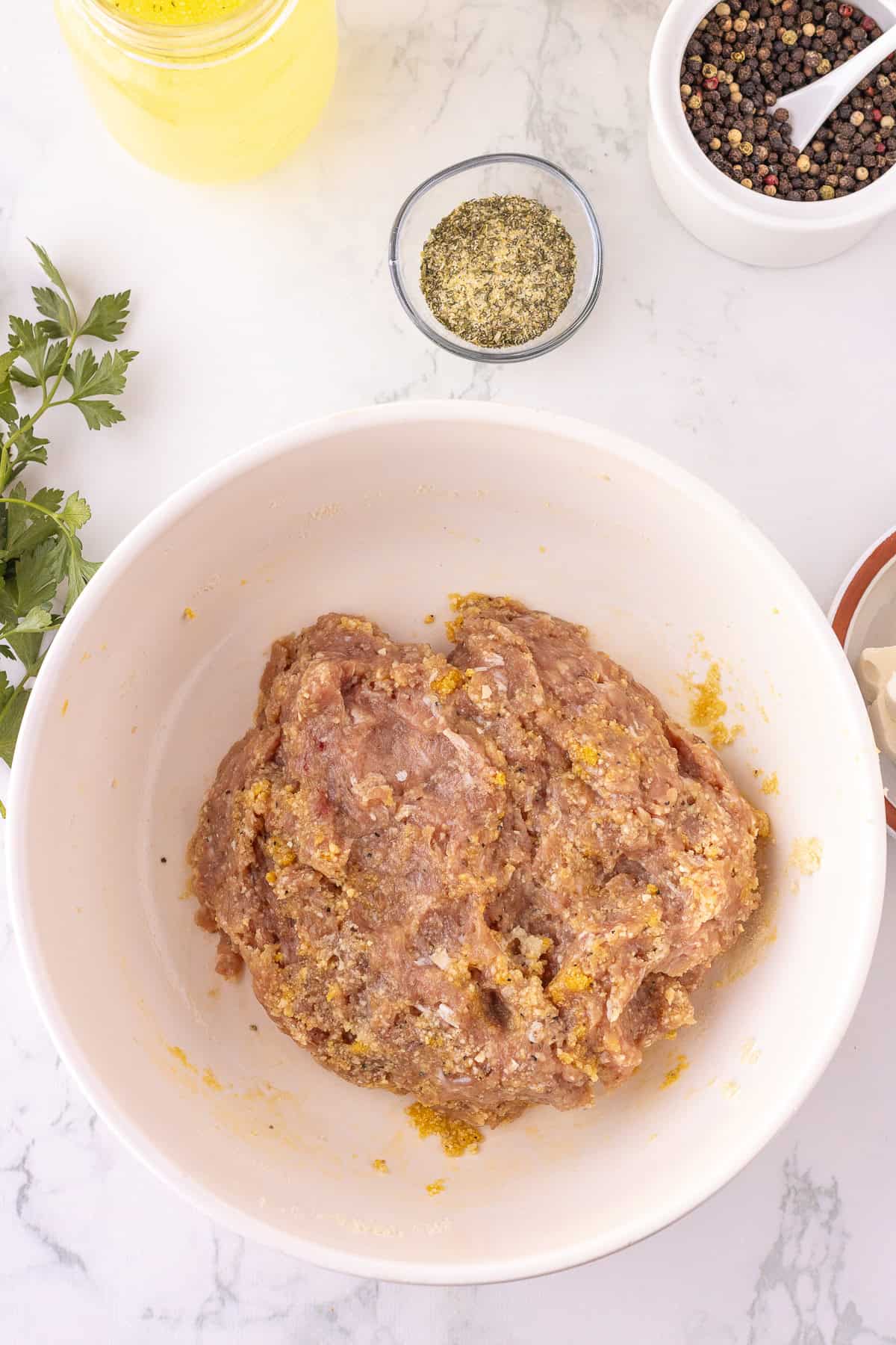 A bowl of seasoned ground meat mixture for Ranch Chicken Meatballs sits on a white surface, surrounded by bowls of spices, peppercorns, and fresh parsley.