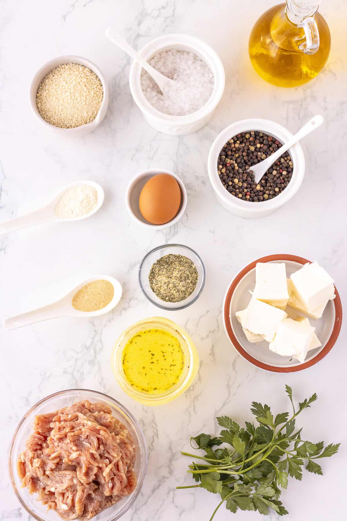 A flat lay of Ranch Chicken Meatballs ingredients: raw ground chicken, an egg, cream cheese, olive oil, spices, breadcrumbs, salt, pepper, broth, Parmesan, ranch seasonnng, and fresh parsley on a white marble surface.