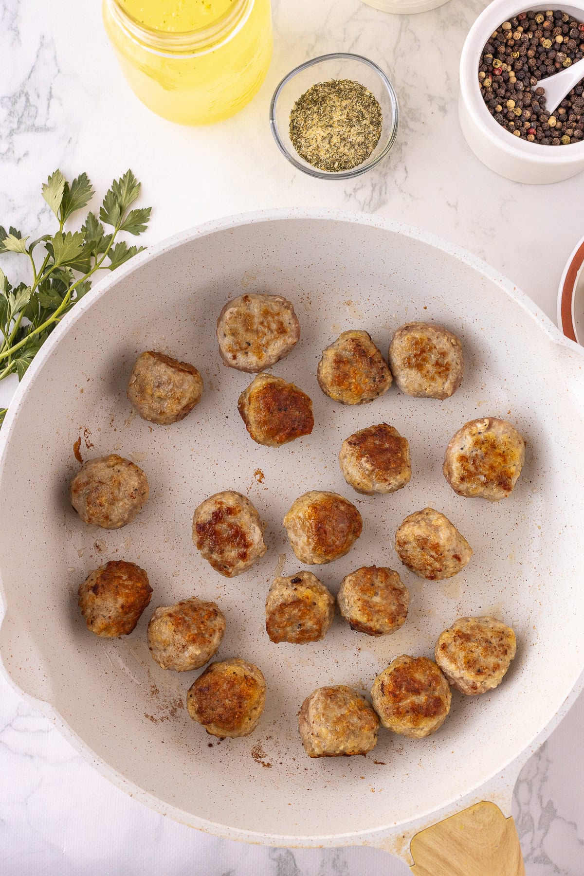 Meatballs are arranged in a white skillet on a marble surface, surrounded by seasoning jars and fresh herbs.