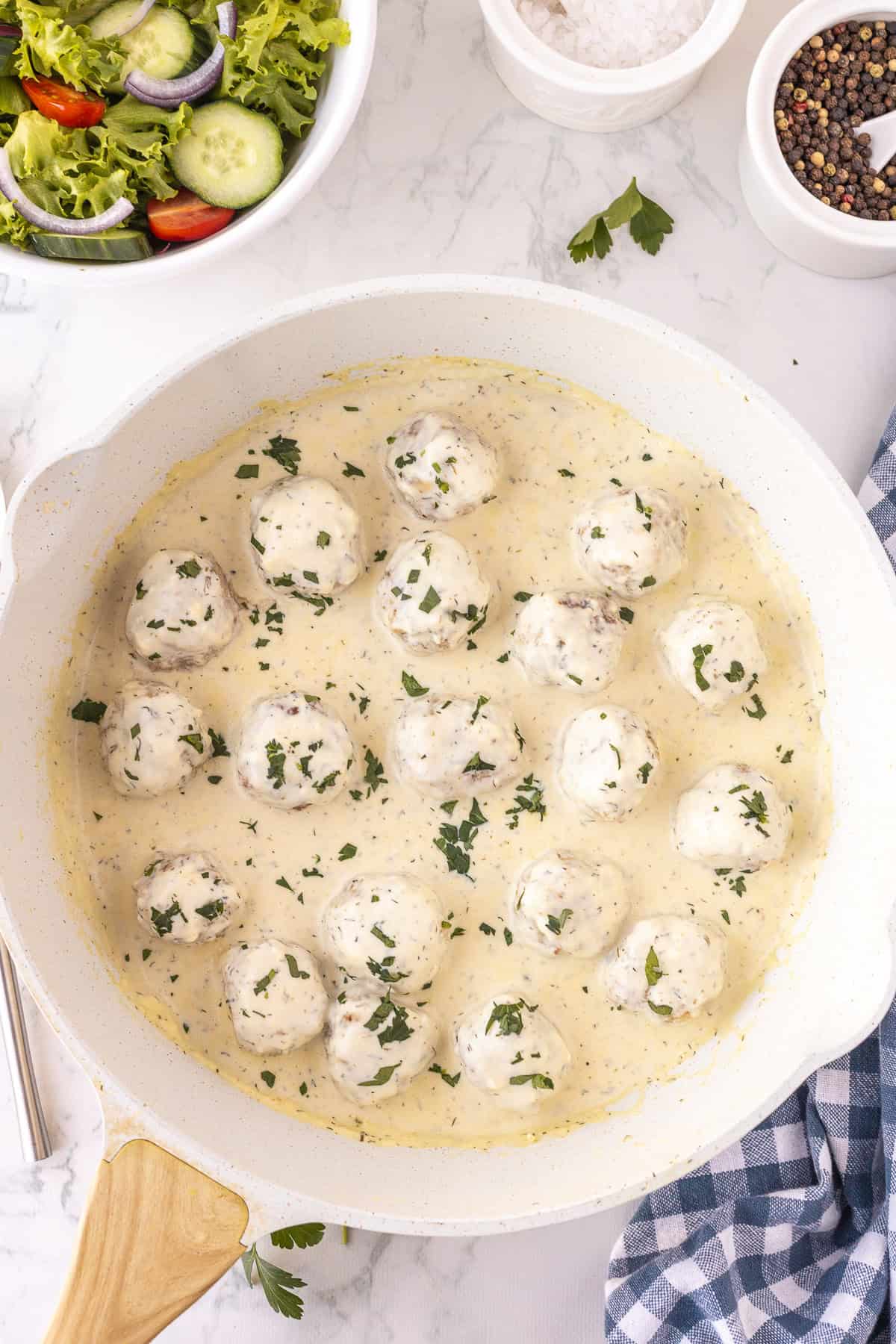 A skillet filled with Ranch Chicken Meatballs in a creamy white sauce, garnished with chopped herbs, sits on a marble countertop next to salad, salt, pepper, and a blue checked napkin.