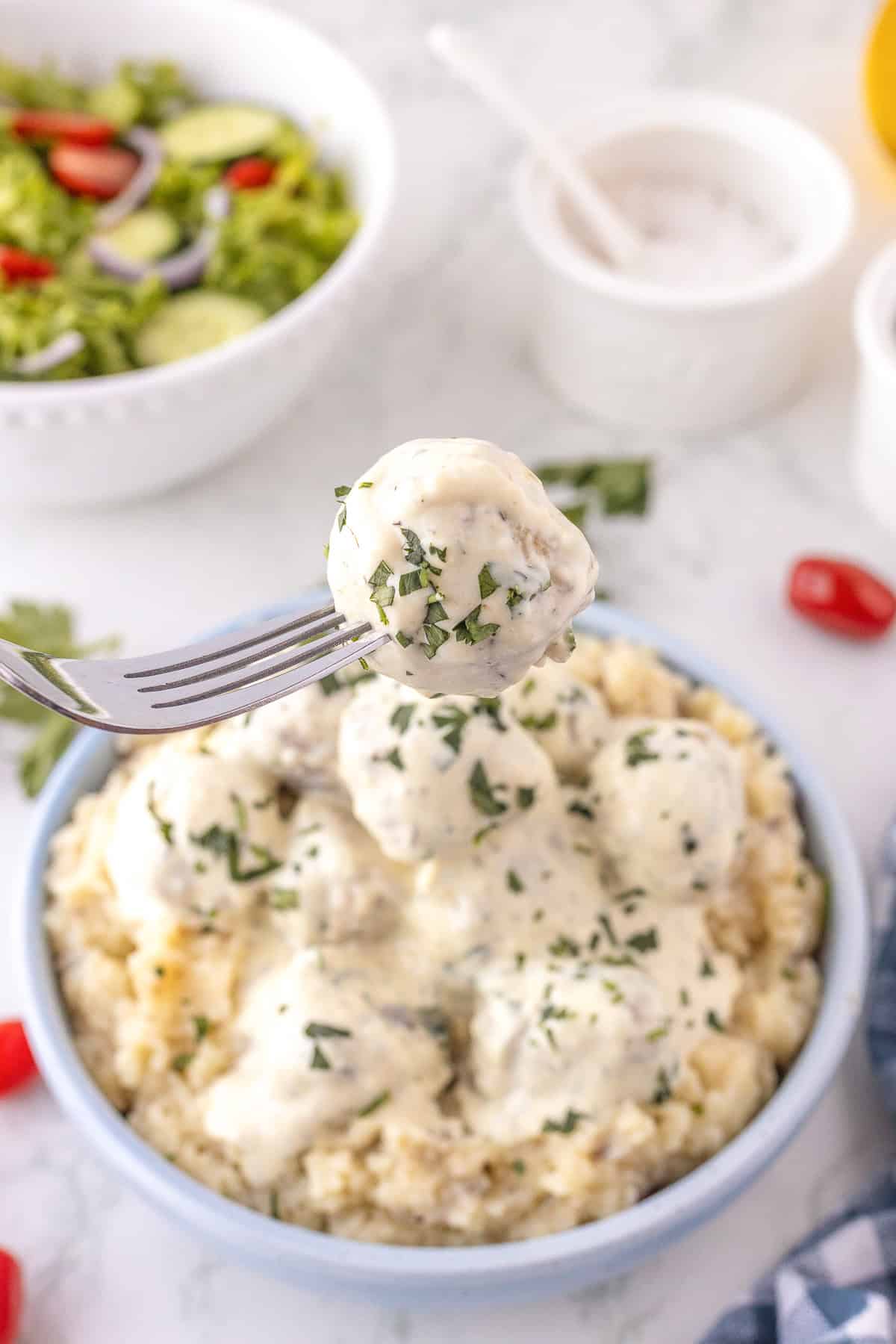 A fork holds a Chicken Meatball with creamy white sauce and herbs above a bowl of meatballs and sauce atop mashed potatoes. A salad and sauce bowl are in the background.