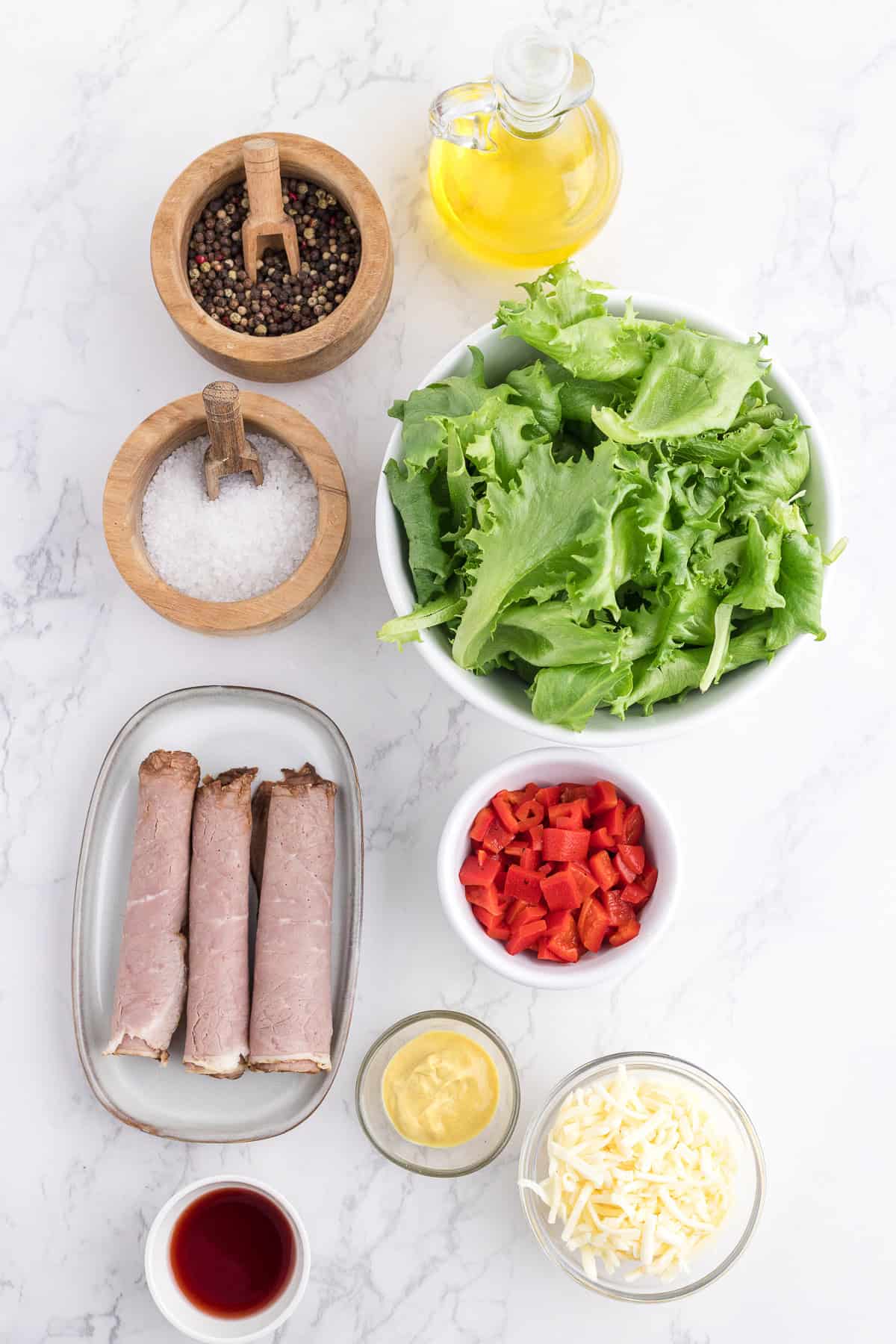 Ingredients for a salad, including green lettuce, rolled roast beef slices, chopped red bell pepper, shredded cheese, mustard, vinegar, oil, salt, and pepper, arranged on a marble surface.