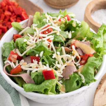 A bowl of leafy green salad with sliced roast beef, shredded cheese, chopped red bell peppers, and dressing.