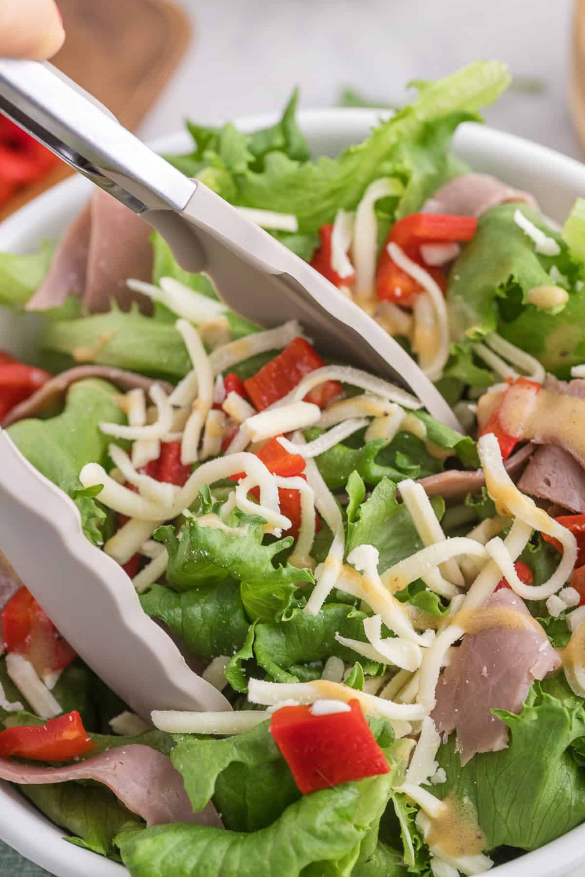 Tongs holding fresh salad with lettuce, red peppers, shredded cheese, slices of roast beef, and creamy dressing in a white bowl.