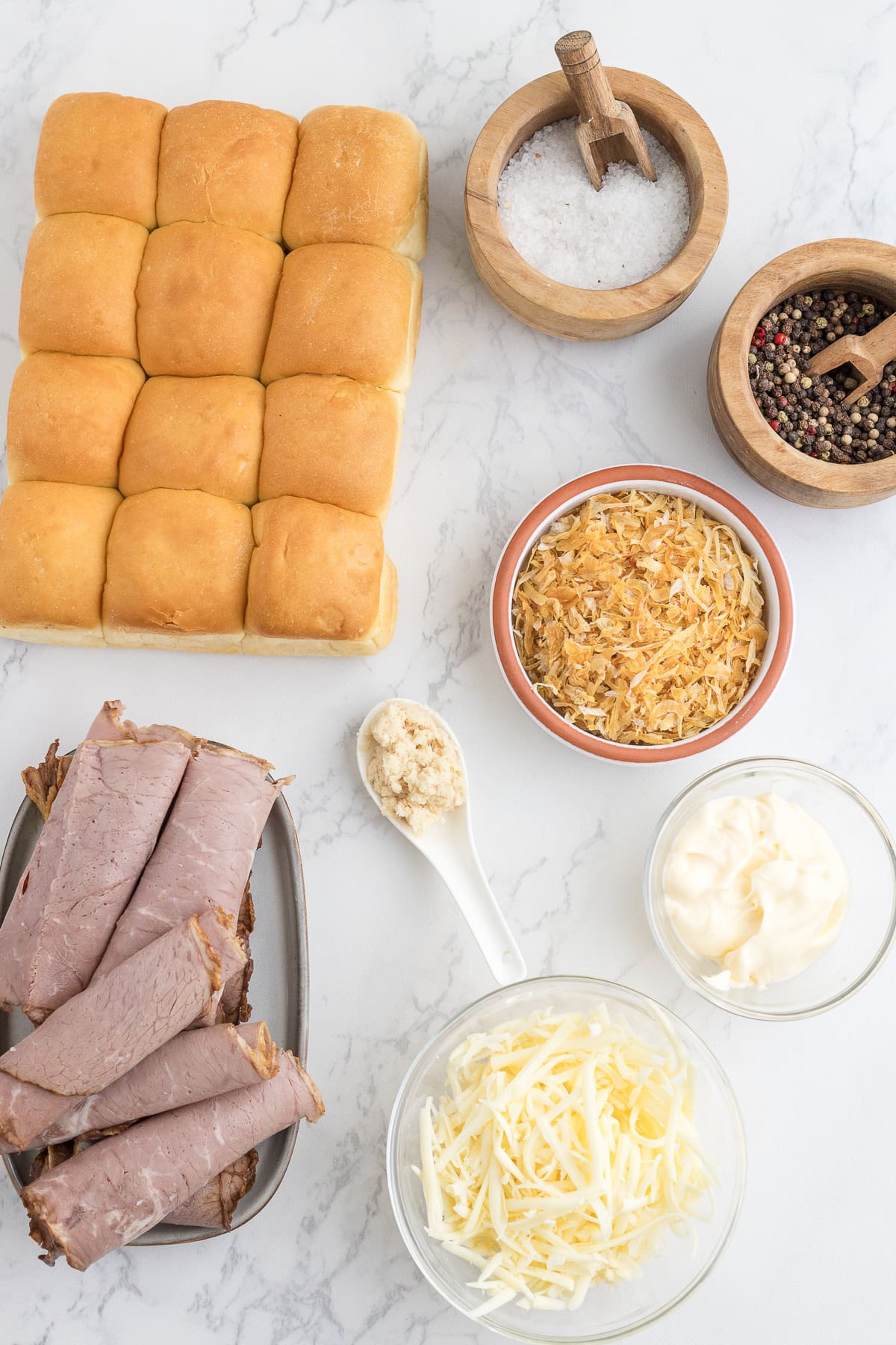 Ingredients for sandwiches arranged on a white surface, including slider rolls, roast beef, shredded cheese, mayonnaise, fried onions, minced garlic, salt, and pepper.