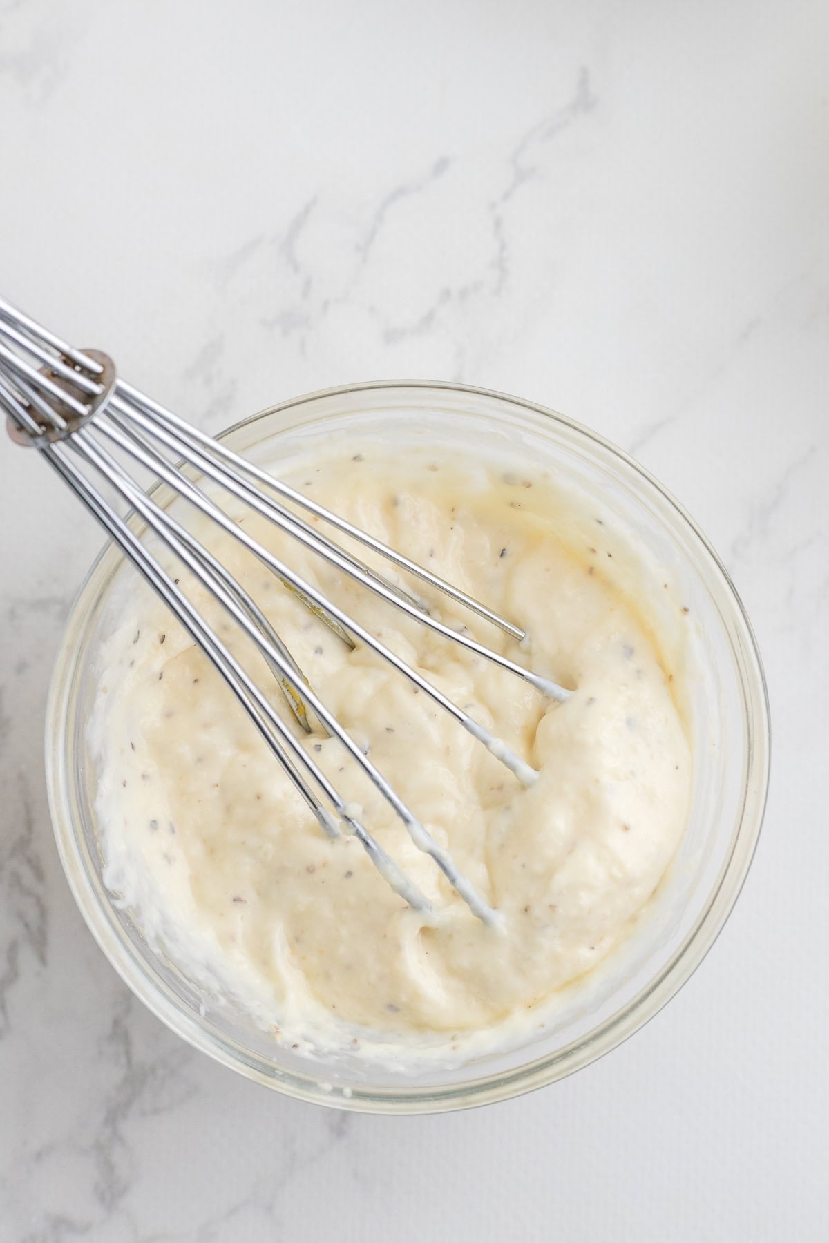 A metal whisk in a glass bowl containing a creamy, seasoned mixture, placed on a white marble surface.