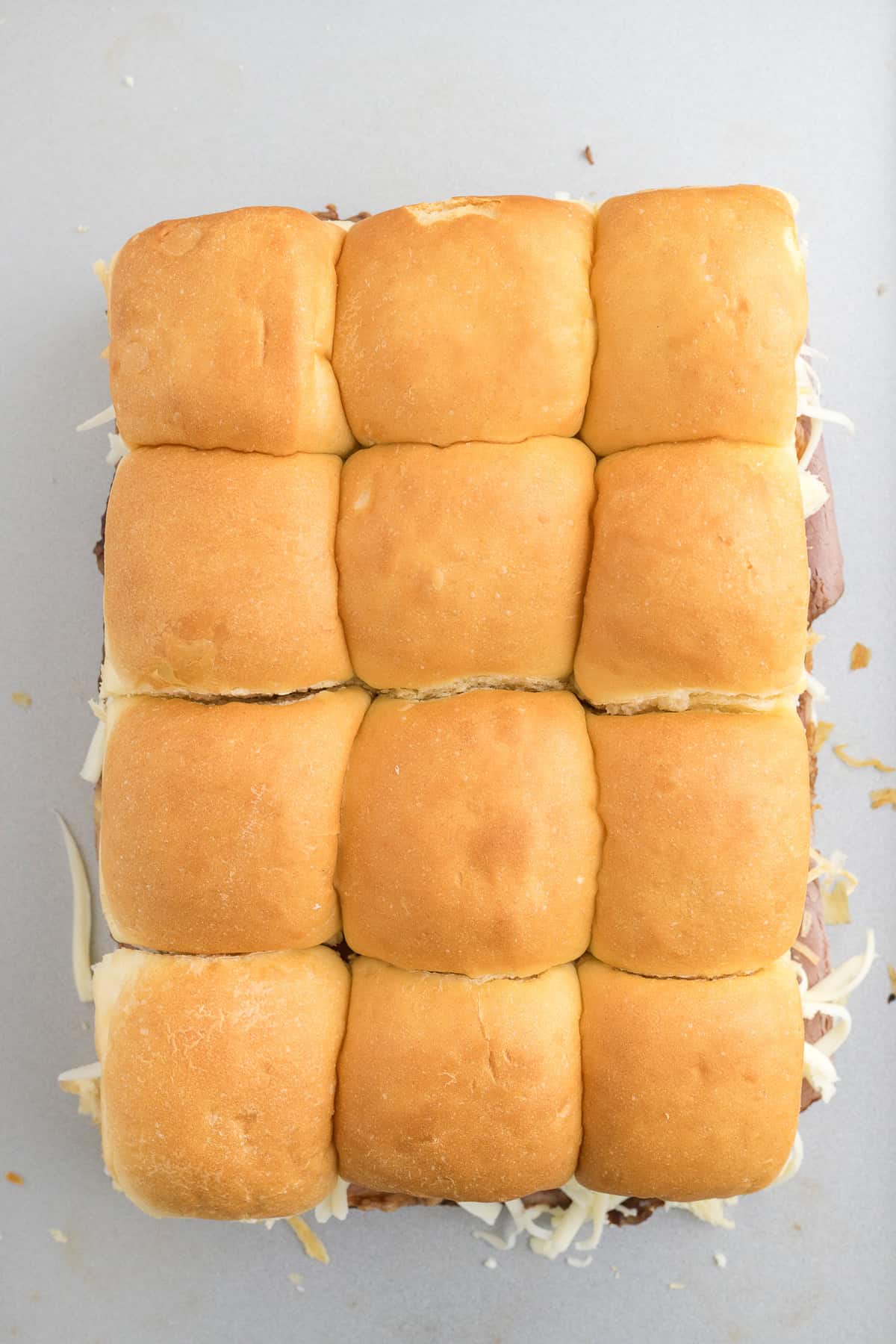 Twelve slider sandwiches with golden-brown buns and visible melted cheese are arranged in a rectangular formation on a light surface.